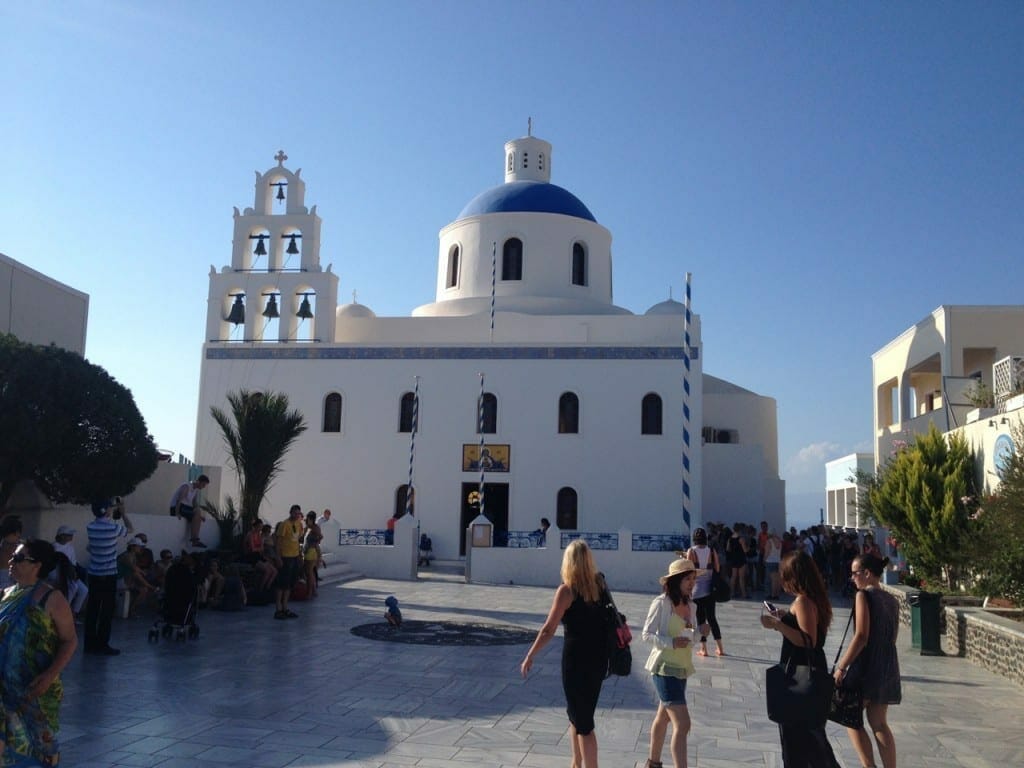people walking at the main square of Oia, Santorini, and a blue-domed church with white walls in the background