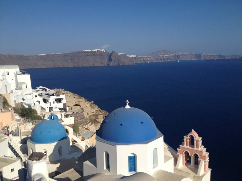 Duas igrejas com cúpulas azul, um campanário salmão, casas caiadas de branco no topo de um penhasco na vila de Oia, Santorini, e ao fundo o azul escuro do Mar Egeu e um longo penhasco