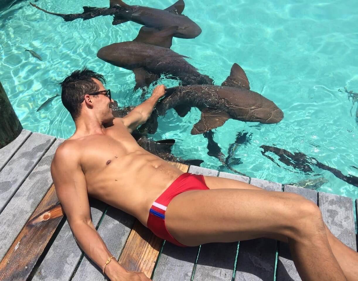 Pericles Rosa wearing sunglasses and a red swim suit  laying down petting sharks at Compass Cay Marina, Bahamas