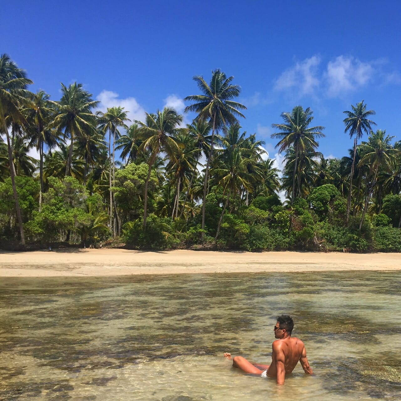 Pericles Rosa sitting on the shallow waters of Moreré Beach on Boipeba Island, with some trees in the backdrop, Bahia, Brazil