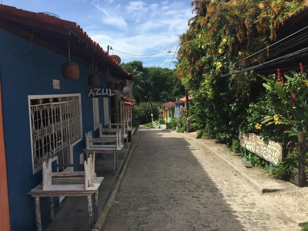 Uma rua no vilarejo de Velha Boipeba, com uma casa azul e janelas brancas de um lado, e árvores do outro lado 