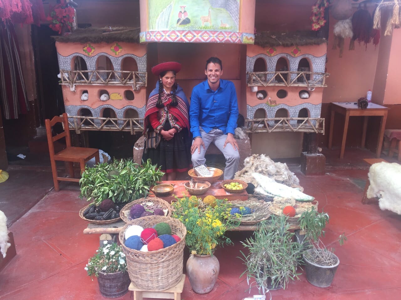 A local Peruvian lady wearing traditional clothes and Pericles Rosa at a weaving workshop in Chinchero, Peru, with  some plants, flowers, cactus, wool, baskets with wool tubes, and bowls with lime, leaves, maize and a tubercle that are used to dye alpacas