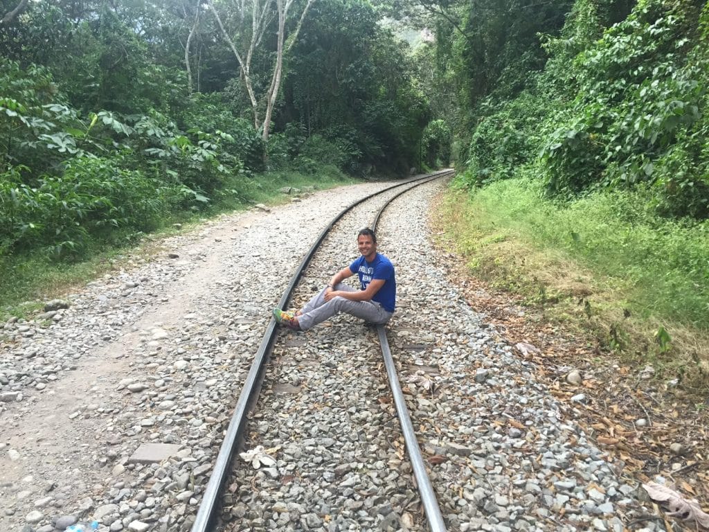 Un hombre con jeans claros, zapatillas de deporte de colores y una camiseta azul, sentado en el
Vías del tren Hidroeléctrica, Aguas Calientes, Perú