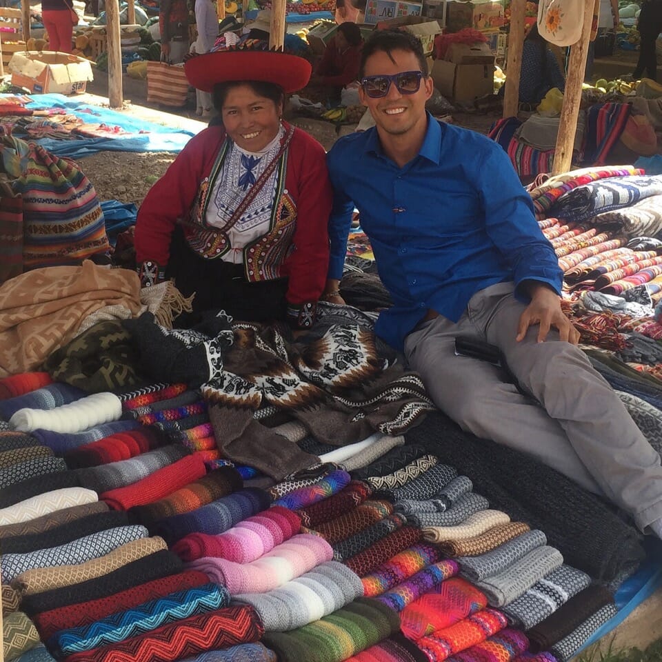 Uma vendedora local vestindo roupas tradicionais e um turista sentado no chão em cima de suéteres no Mercado Chinchero