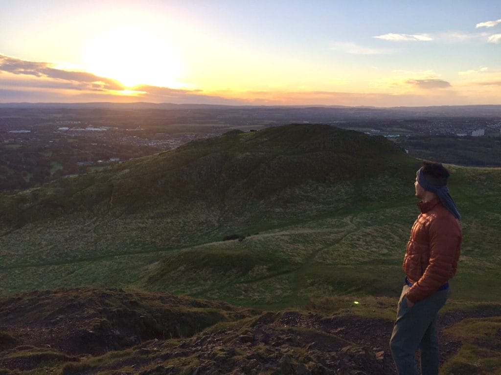 A man wearing an orange jacket standing at the top of Arthur's seat watching the sunrise.