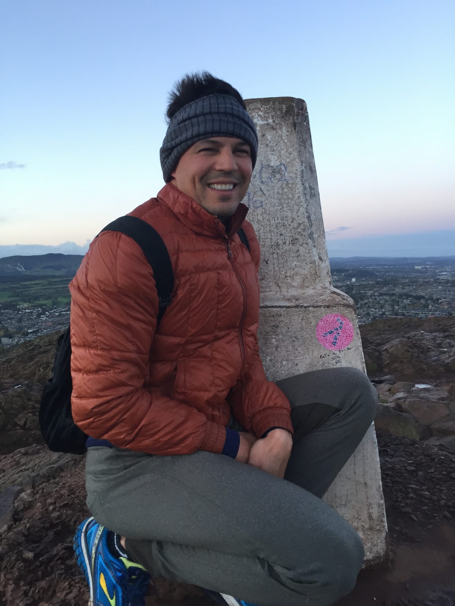 A man wearing an orange jacket and grey trousers on the top of Arthur's Seat, Edinburgh, Scotland