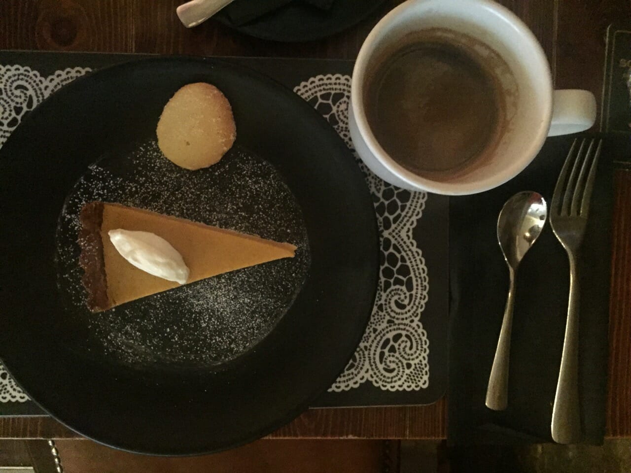 a piece of pumpkin pie and a cookie served on a black plate and a white cup of coffee on the top of a white place mat 