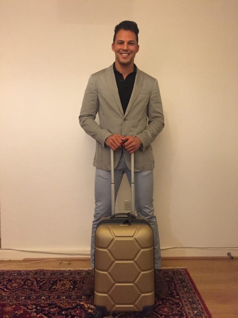 A man wearing a grey summer blazer, a black polo and a light-blue trouser holding a golden carry-on suitcase