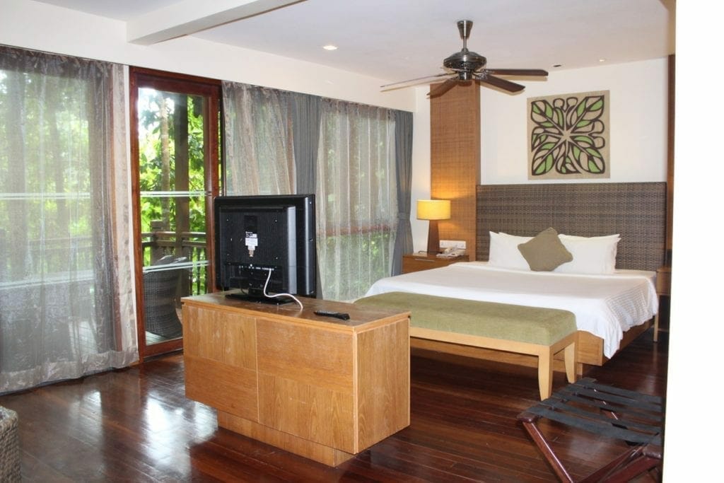 estudio bosque tropical en el Berjaya Langawi Resort con muebles y TV LCD, pisos de madera, puertas de vidrio y ventanas con cortinas, cama king size, marco, ventilador de techo, lámpara y mesita de noche