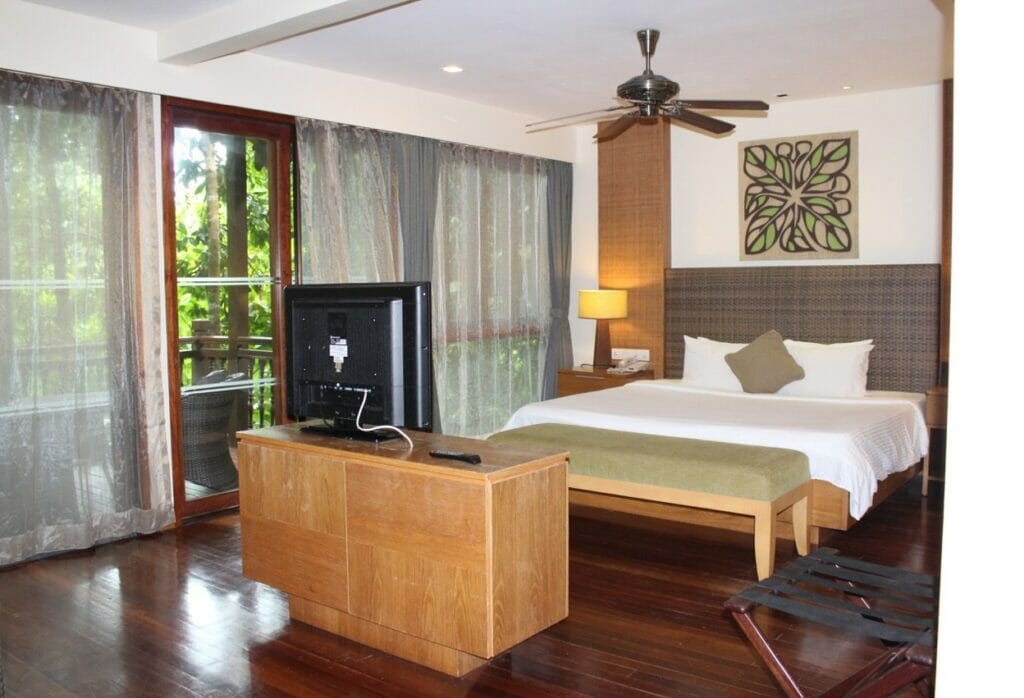 Rainforest bedroom of Berjaya Langkawi Resort finely decorated with double bed, pillows, TV, fan, window glasses adn wood floor.