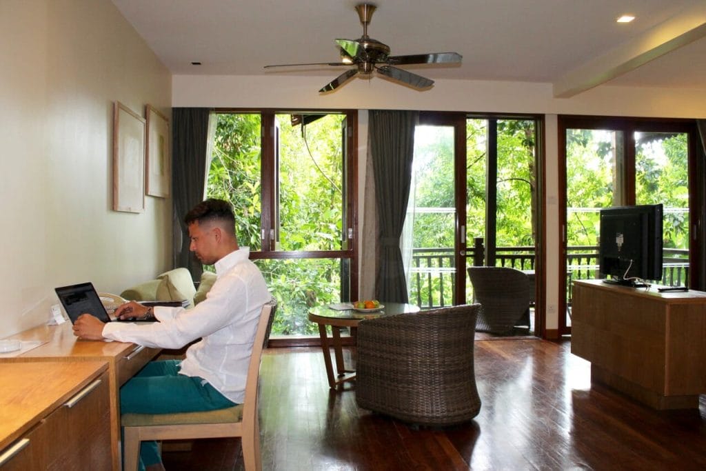 Hombre vestido con una camisa blanca y pantalones verdes trabajando en la computadora en una habitación con pisos de madera, muebles, televisión LCD, ventilador de techo, puertas y ventanas de vidrio