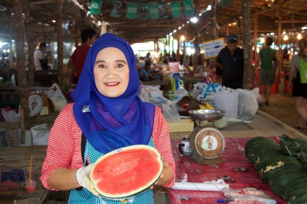 A woman wearing a blue headscarf and white blouse with red dots holding half of a watermelon at Ao Nang's local market