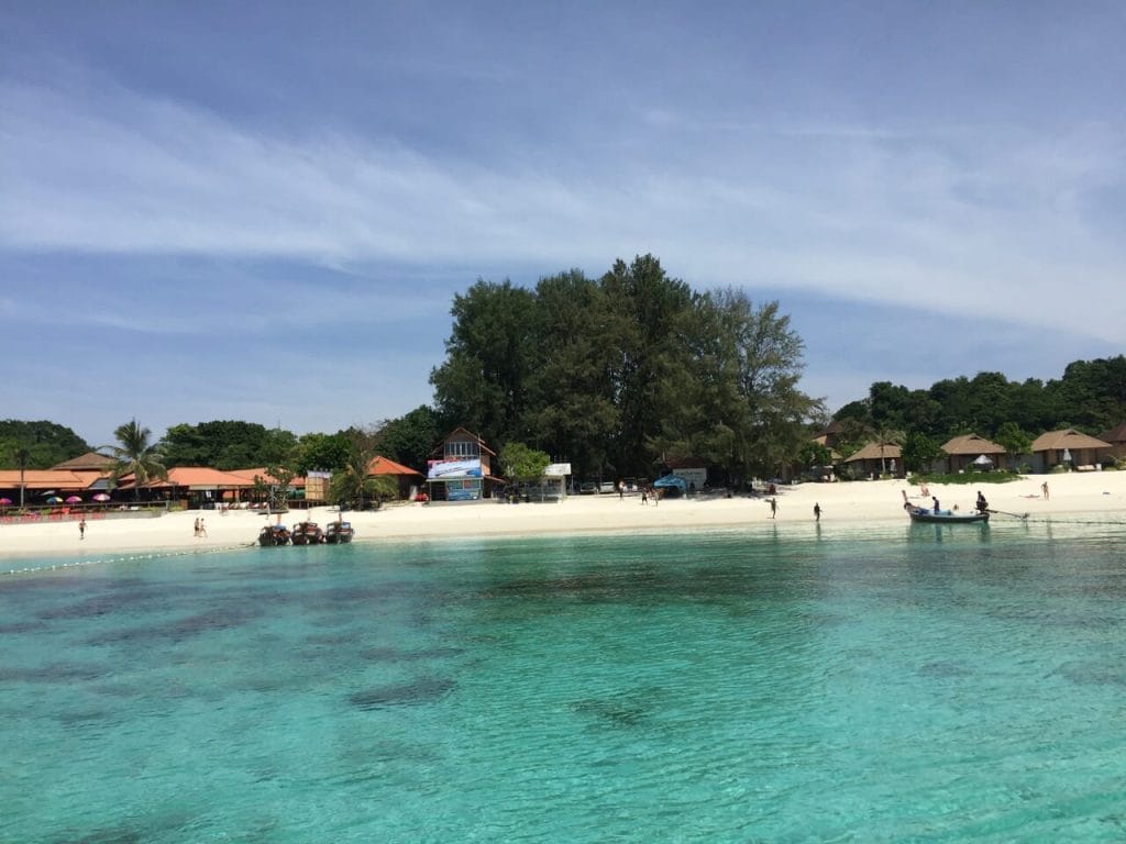 The crystal turquoise water of Pattaya Beach, backed up by some trees, hotels and traditional long-tail Thai boats on the water, Koh Lipe, Thailand