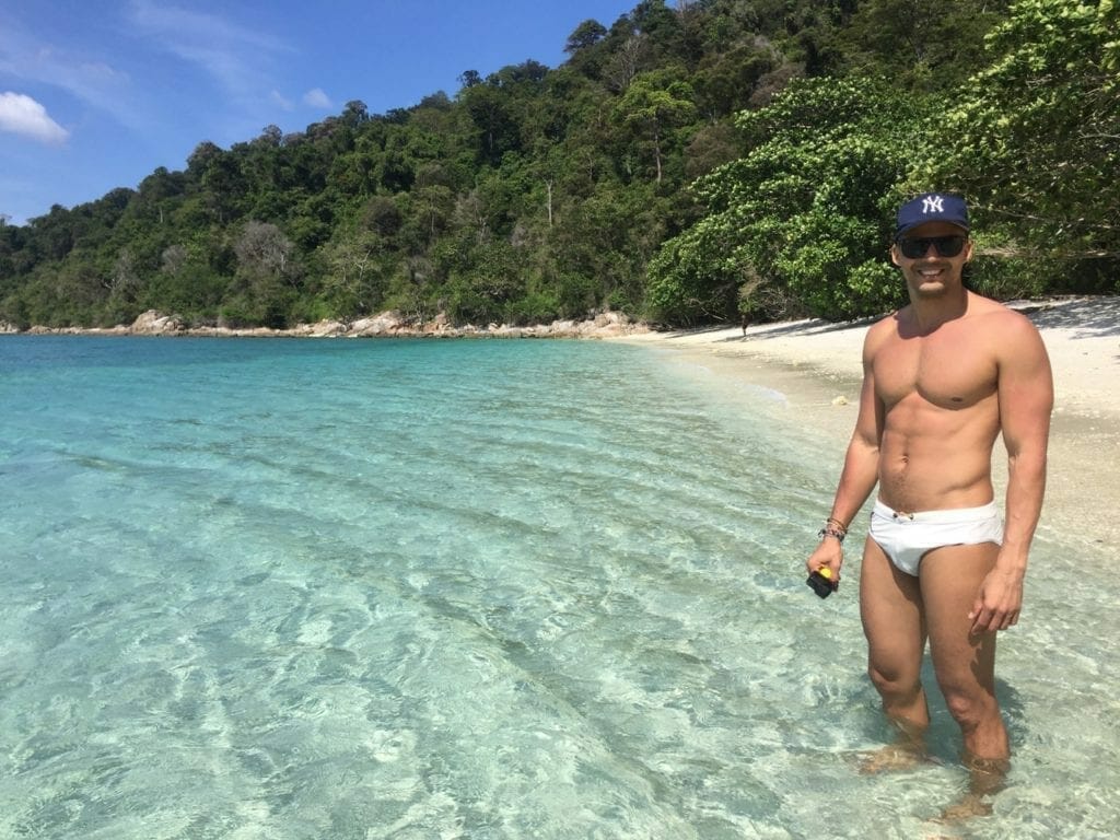 Un hombre con una gorra azul, gafas de sol y bañador blanco de pie en una isla desierta en el sur de Tailandia