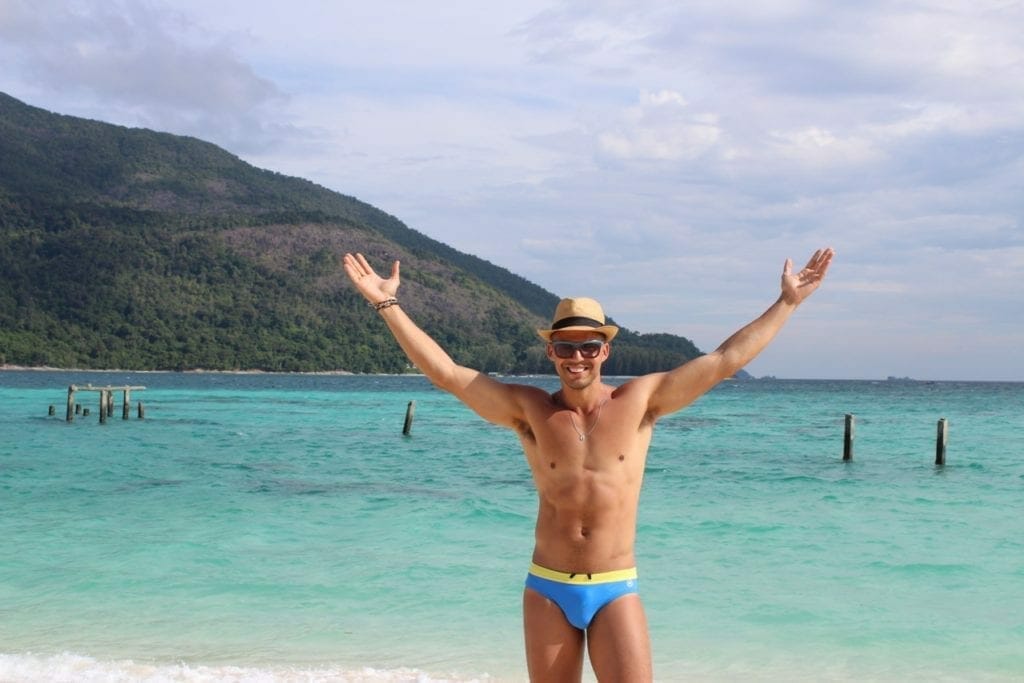 Pericles Rosa con un sombrero marron, gafas de sol y un bañador azul y amarillo con los brazos abiertos en Sunrise Beach, Koh Lipe, Tailandia 