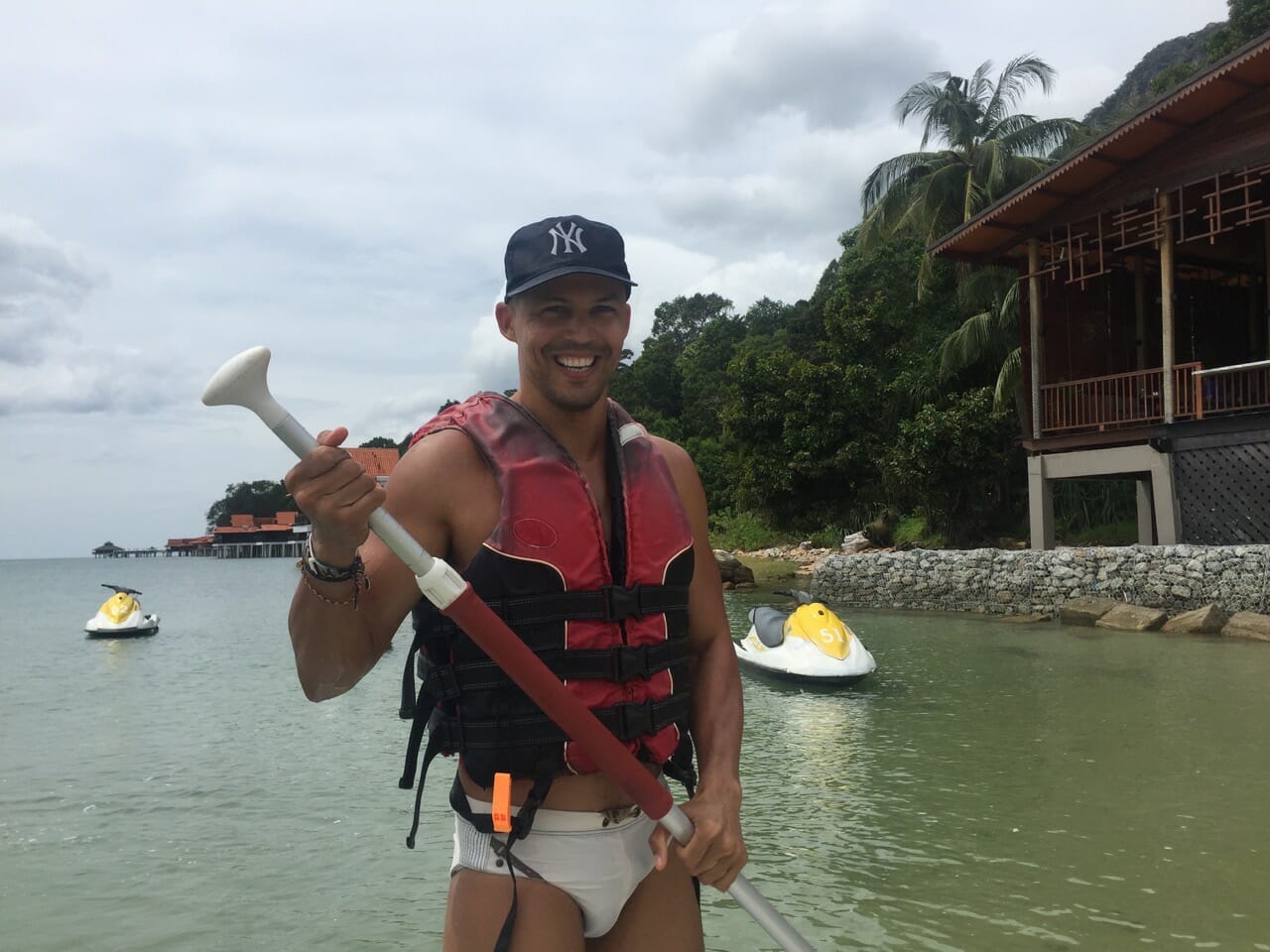 a man wearing a hat, red life jacket and white speedo doing stand up paddle boarding on the beach with tow jet skies on the water