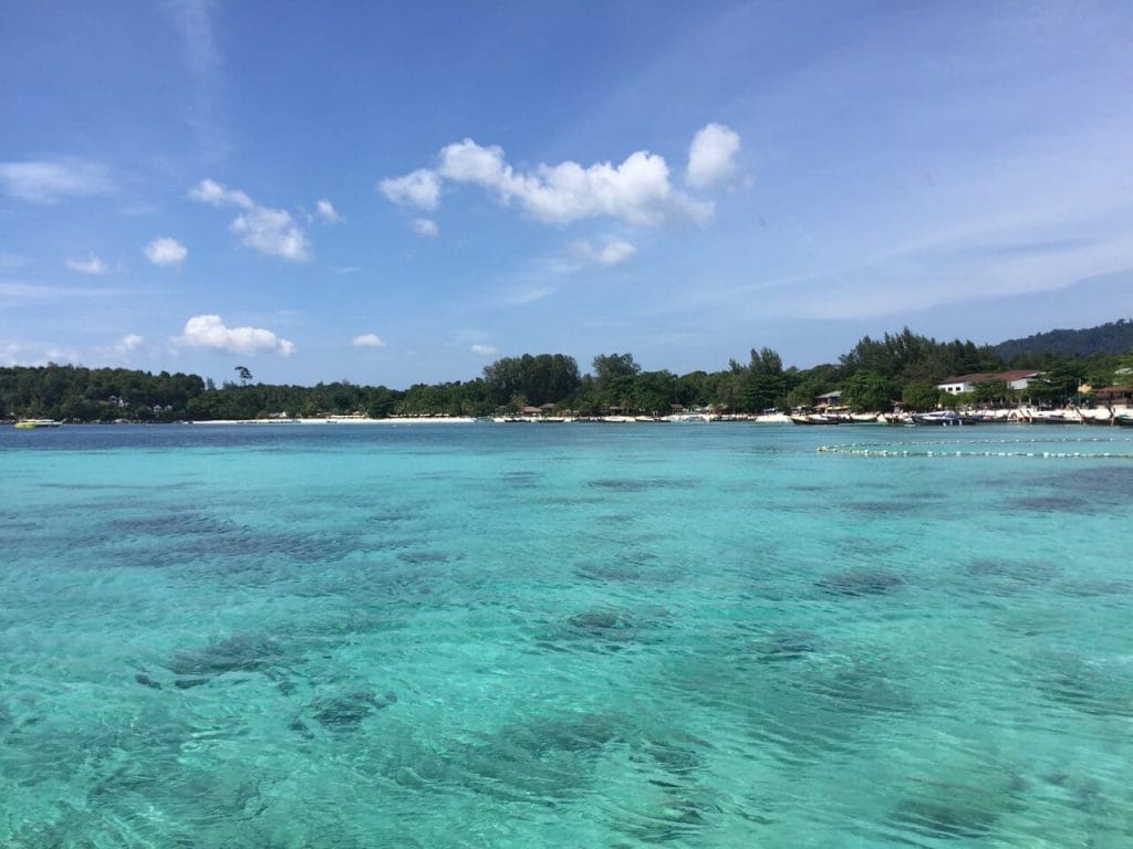 The crystal-clear turquoise water of Pattaya Beach in Koh Lipe, backed-up by vegetation, long-tail boats and blue sky 