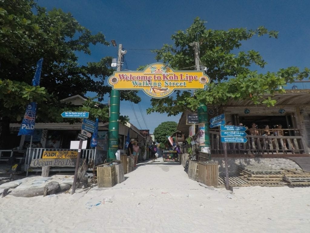 A rua principal da ilha de Ko Lipe, Tailândia, na entrada tem areai da praia, bares de madeira, árvores e um placa amarela onde se lê 