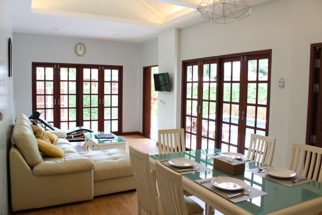 the living room of a villa at Alisea Pool Villas with a dinner table, chairs, sofa, pillows, a small tv on the wall and glass doors with wood frames