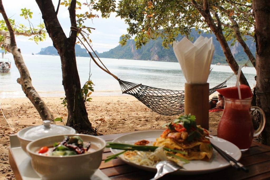 Mesa para el almuerzo en Adamana Beach Club, Aonang, con platos, una jarra de jugo de sandía, una hamaca y al fondo el mar y las montañas de piedra caliza cubiertas de vegetación