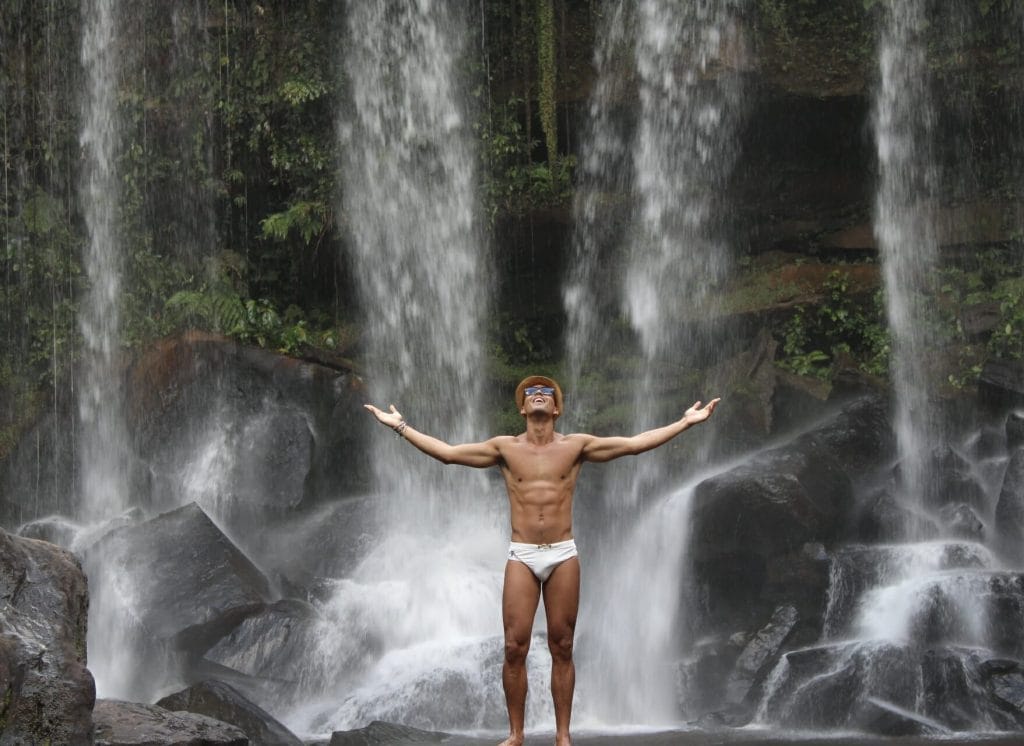 Pericles Rosa wearing a hat, blue sunglasses and a white speed with his arms wide open in Kulen Mountain, Camboja