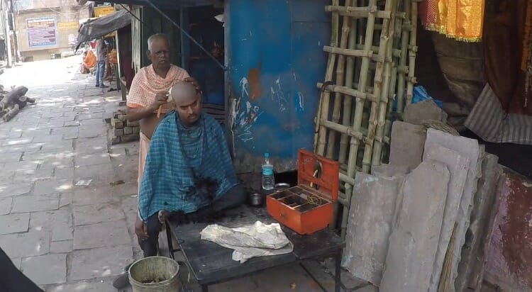 um homem vestindo uma capa azul e outro homem usando uma roupa salmão cortando o cabelo dele