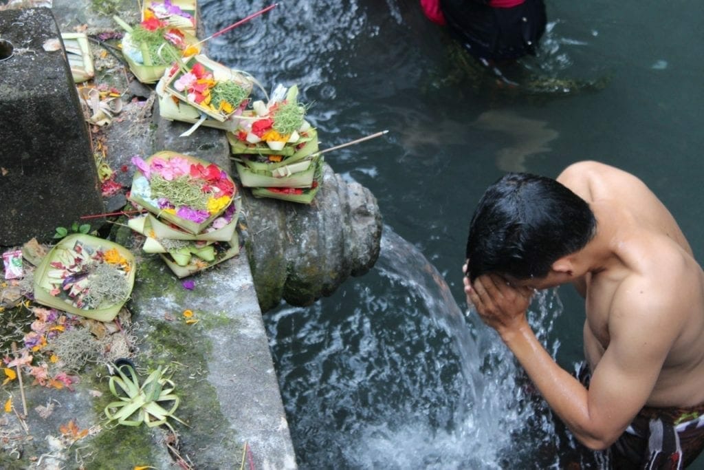 Un hombre en pantalones cortos y sin camisa en el agua rezando junto a algunas ofrendas en Tirta Empul, Bali.