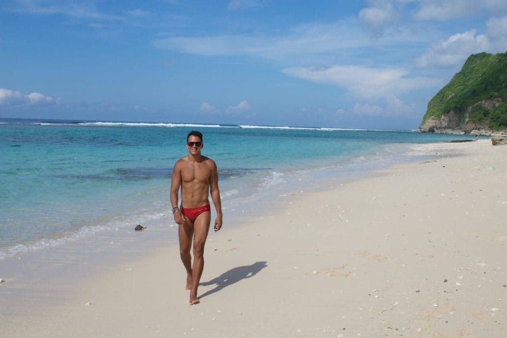 Péricles Rosa vestindo uma sunga vermelha e óculos escuros caminhando na praia de Karma, em Bali, e as águas cristalinas e o penhasco marítimo coberto de vegetação ao fundo