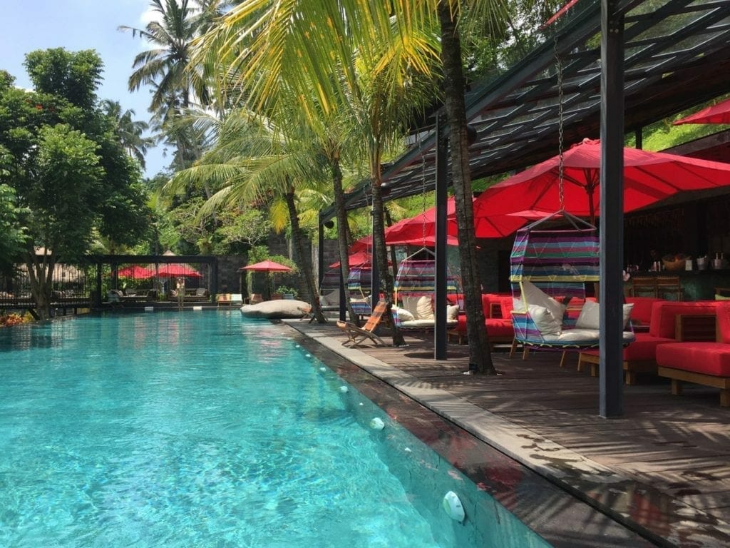 Uma linda piscina, alguns guarda-sóis vermelhos, sofás vermelhos e cadeiras coloridas rodeadas por uma vegetação luxuriante em Jungle Fish, Bali