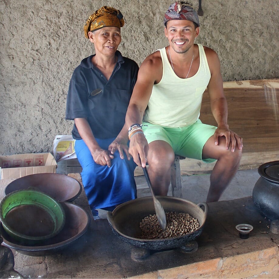 Péricles Rosa usando um lenço indonésio na cabeça, uma regata amarela e um short verde mexendo alguns grãos de café em uma panela com uma mulher indonésia vestindo uma camisa pólo azul, saia azul e um lenço na cabeça na Lumbung Sari House of Coffee em Bali