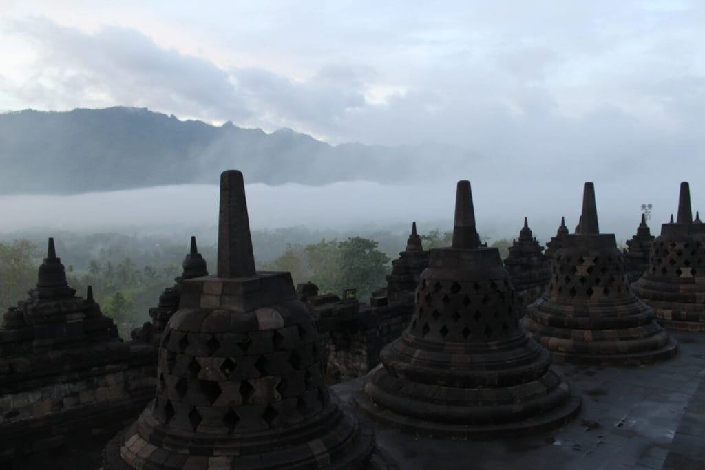 As stupas pretas e acastanhadas, as árvores e a neblina compõem um lindo cenário durante o amanhecer no Templo de Borobudur