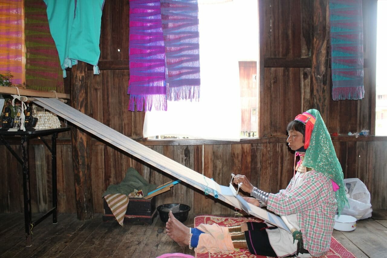 A long neck women working on a loom in a wood stilt-house of a weaving factory with a window and some scarves hanging on the ceiling 