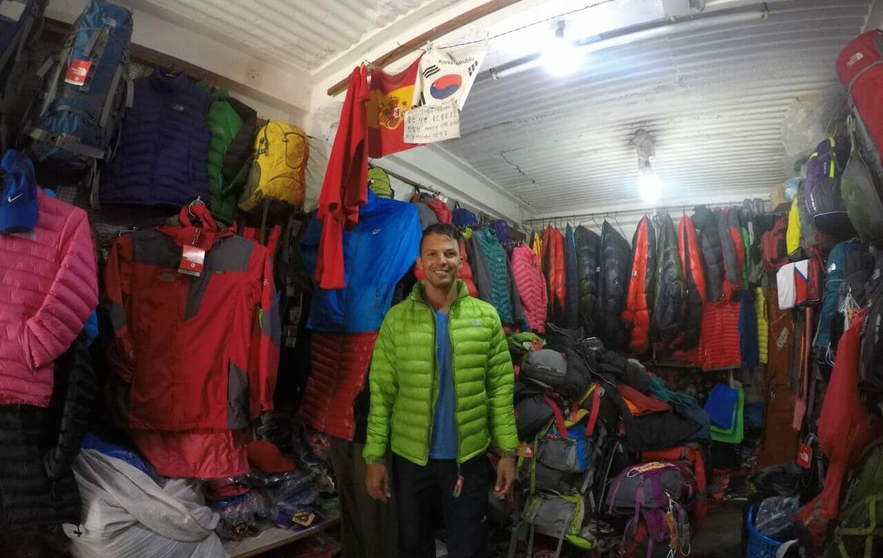 Pericles Rosa wearing a light-green jacket, blue t-shirt and dark blue trouser in the middle a store in Pokhara, Nepal, with many jackets of different colours hanged on the walls