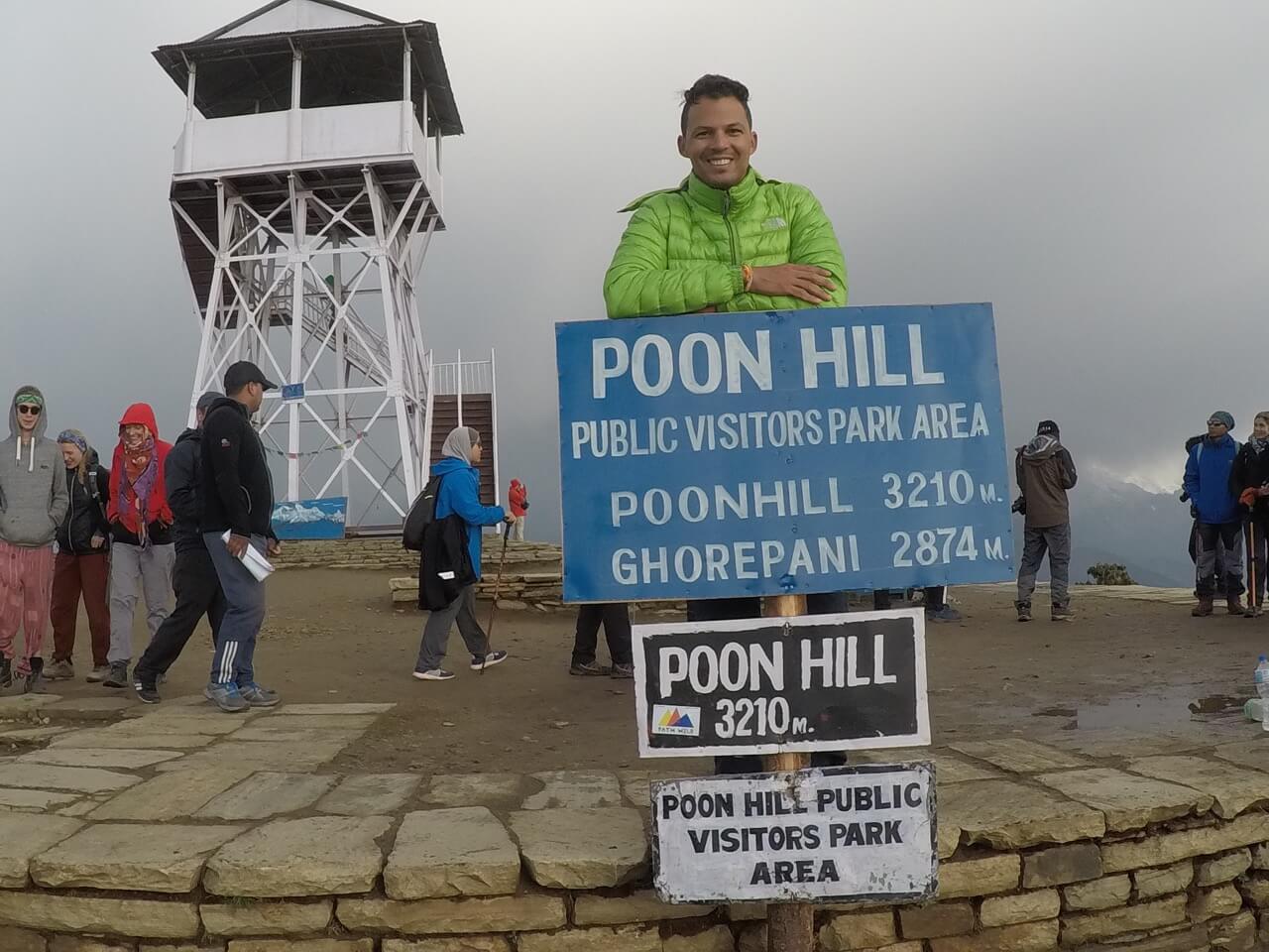 Pericles Rosa wearing a light-green jacket and some other people at Poon Hill Public Visitors Park Area on the top of Poon Hill Mountain leaning on a blue sign and a white tower that servers as a viewpoint in the background