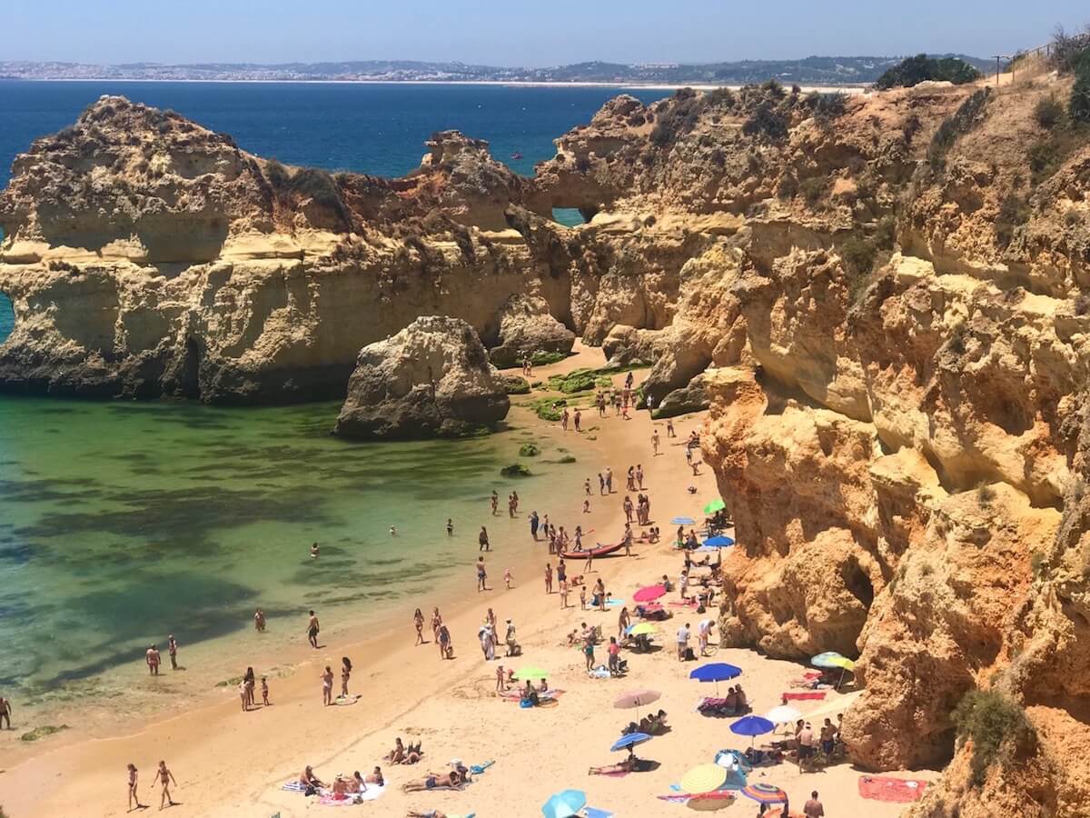 Gente paseando y tomando el sol en Prainha, Alvor, Portimão, Portugal, que está rodeada de enormes acantilados de piedra caliza de color ocre y tiene aguas verdes cristalinas.