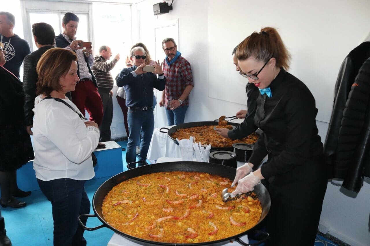 dos enormes ollas de paella, una comida hecha con arroz, pollo y camarones, con una mujer vestida de negro sirviendo una ración a otra mujer mientras dos hombres las fotografían