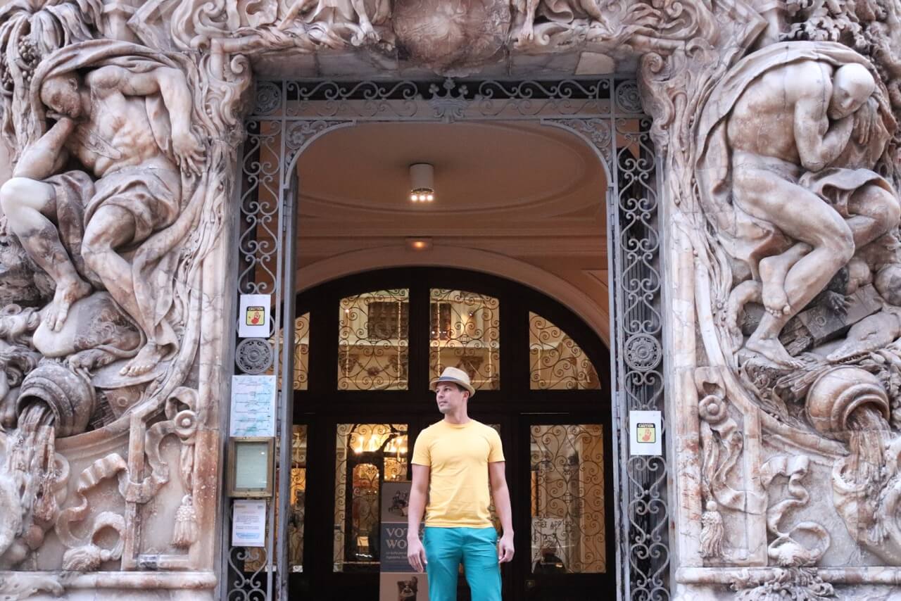 um homem usando um chapéu, camiseta amarela e calça verde saindo do Museu de Cerâmica de Valência que tem uma fachada impressionante de mármore esculpido com potes de água e um homem em cada lado