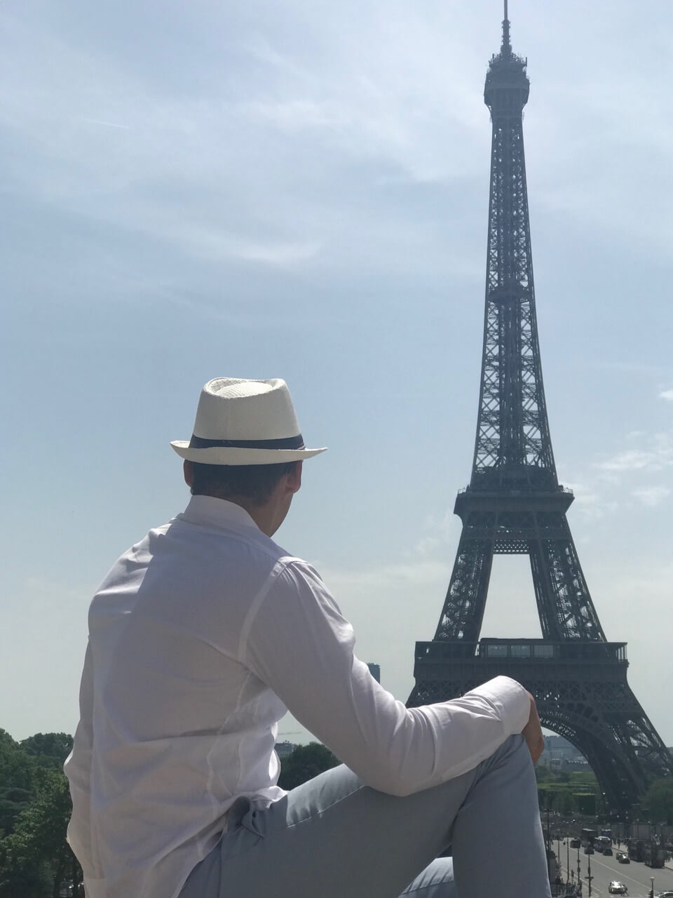 un hombre con una camisa blanca, sombrero blanco y pantalón celeste sentado en El Trocadéro, Paris, admirando la Torre Eiffel
