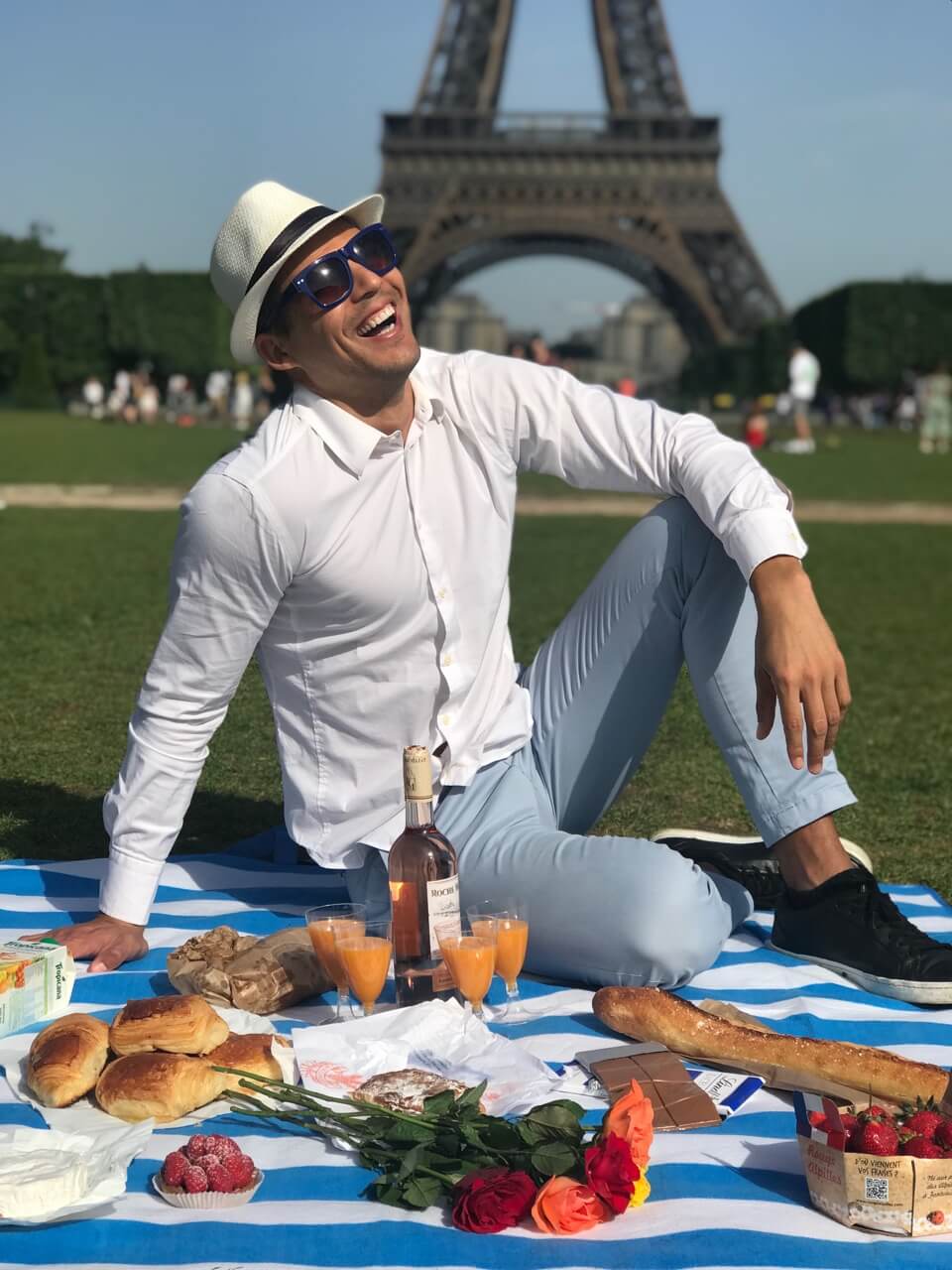 Um homem vestindo uma calça azul clara, uma camisa branca, um chapéu branco e um óculos azul fazendo um piquenique no Camps de Mars, em Paris, com a Torre Eiffel de fundo