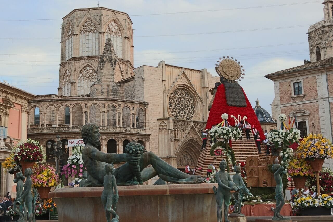Plaza de la Virgen, Valencia, con una fuente y varias estatuas y al fondo una enorme imagen de la Virgen María y parte de la catedral de la ciudad.