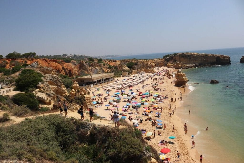 Praia de São Rafael, Albufeira, que está rodeada de acantilados de color naranja con arena dorada y agua turquesa cristalina, con muchas sillas y sombrillas, un restaurante y gente caminando sobre la arena y tomando fotografías desde su mirador