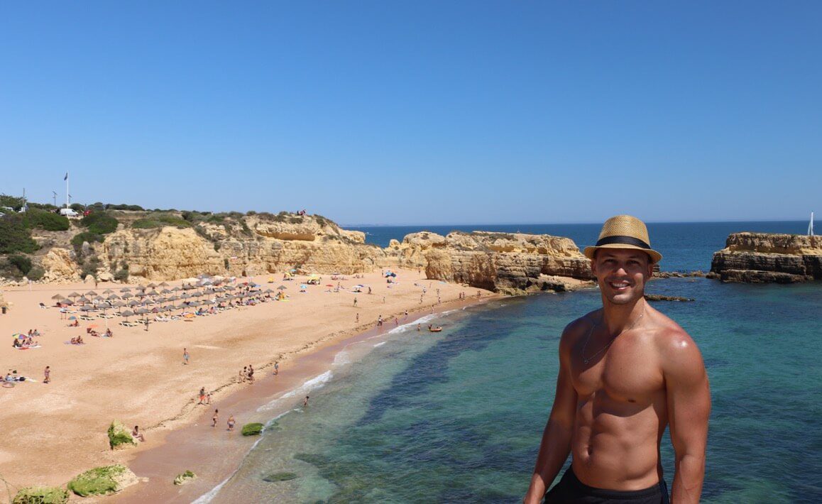 Pericles Rosa usando um chapéu marrom e um short preto posando para uma foto em cima de uma falésia com a Praia do Castelo ao fundo, Albufeira, Portugal