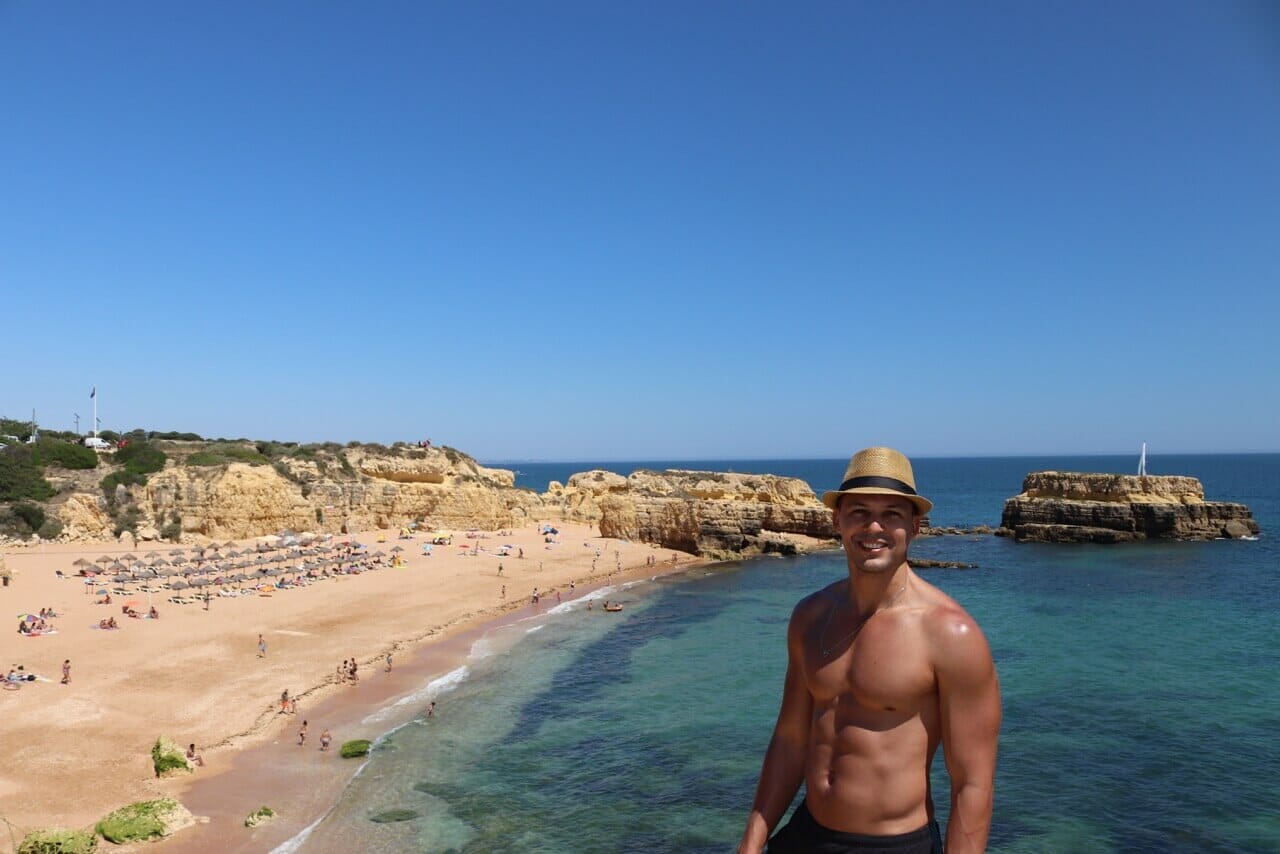 Un chico de sombrero marron y short negro posando para una foto en cima de un acantilado con el mar al fondo en la Playa del Castillo, Albufeira, Portugal