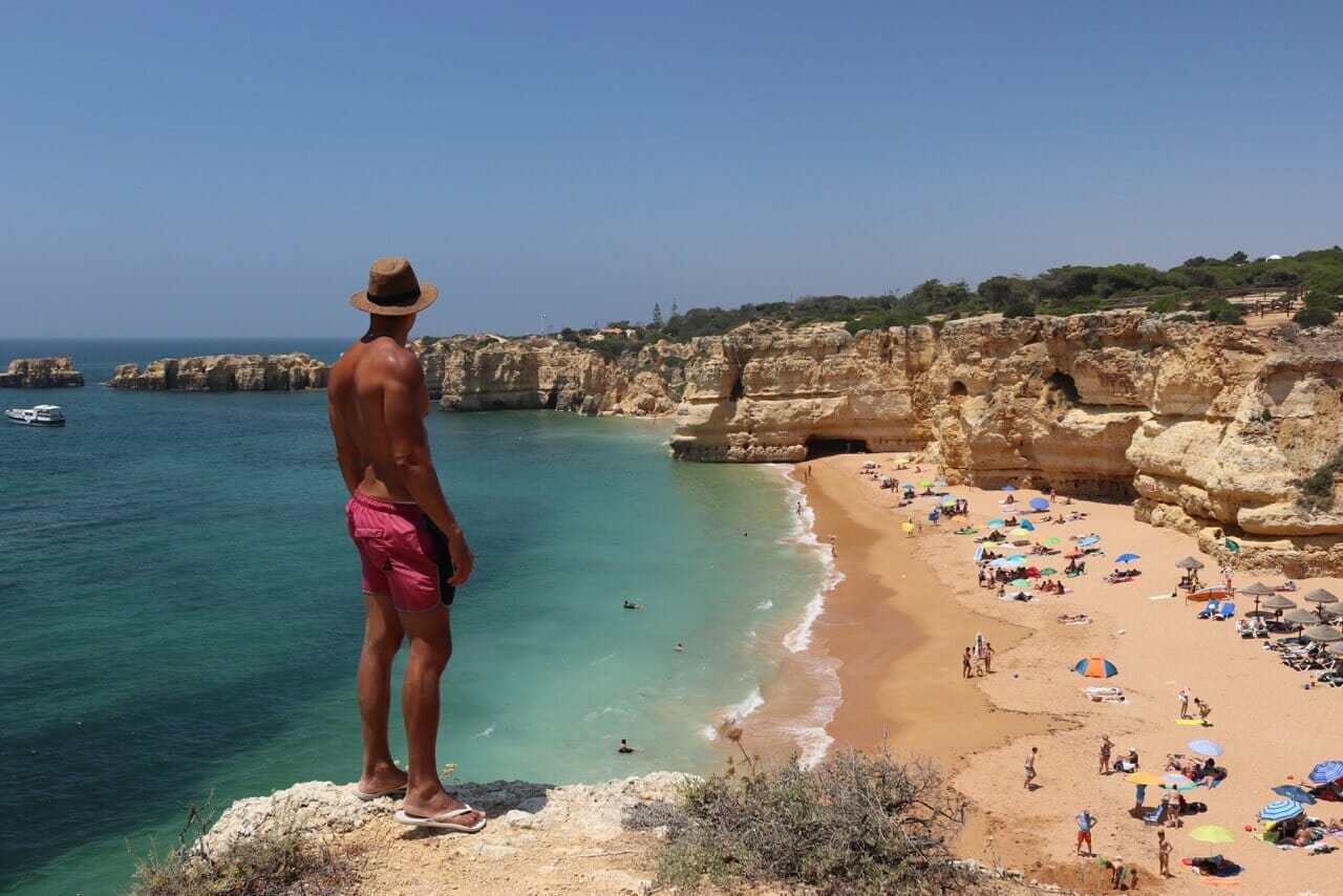 Un hombre con um sombrero y un short rojo an el top de un acantilado mirando la playa en Praia da Coelha, Albufeira, Portugal