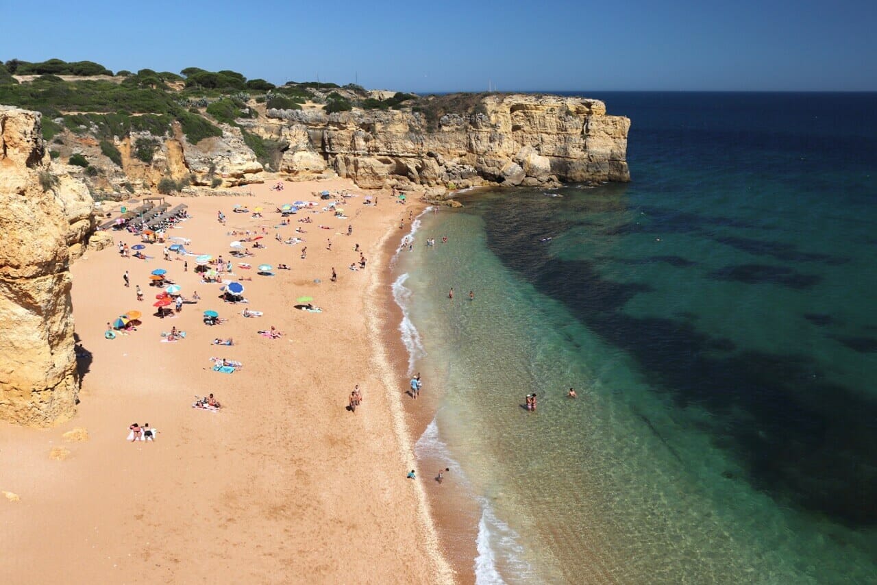 El extraordinario paisaje de Praia da Coelha, que está rodeada de acantilados de piedra caliza amarilla, tiene arena de color ámbar suave y agua azul translúcida con gente tomando el sol, paseando por la playa y en el agua