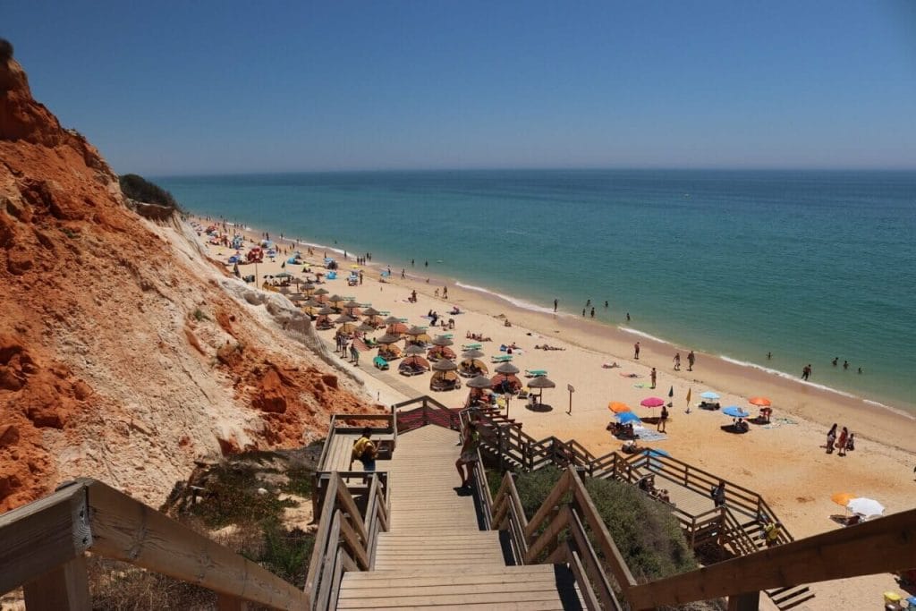 Una escalera de madera que da acceso a la Praia da Falésia en Albufeira, con un acantilado rojo en el lado izquierdo, la playa con algunas sombrillas y gente en el agua