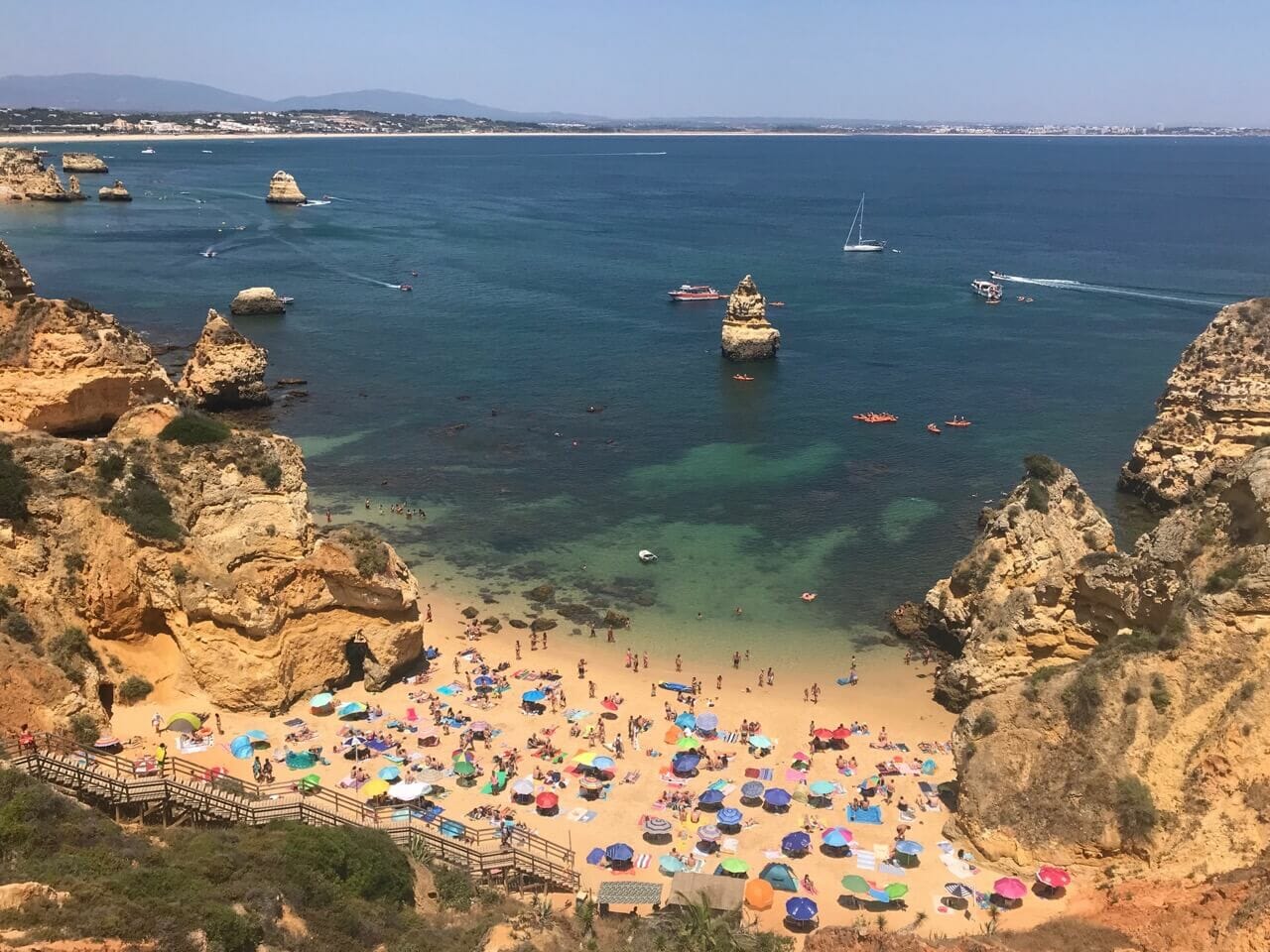 Praia do Camilo, Lagos, enveloped by orange, yellow and red cliffs, with many umbrellas on the amber sand, boats and kayaks sailing on the crystalline green-blue water, and a long wooden staircase. 