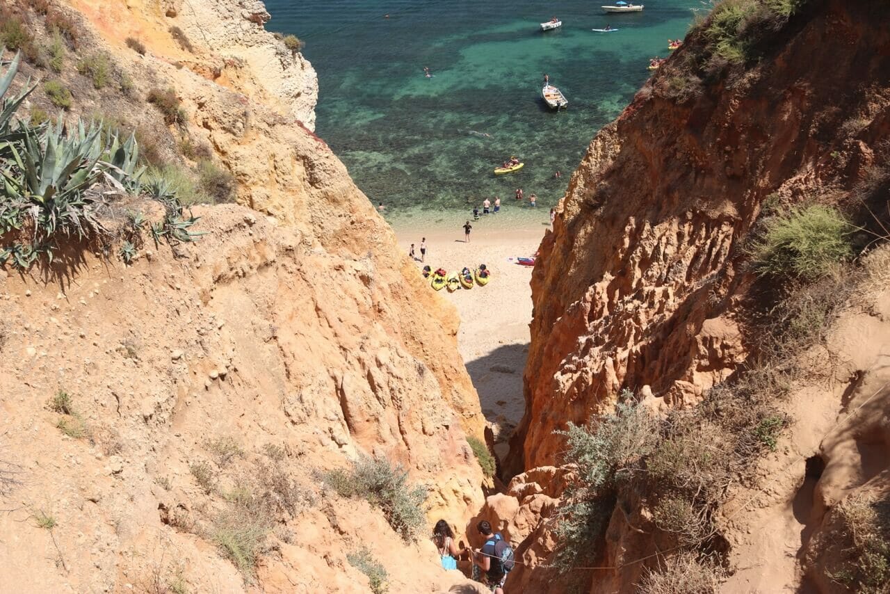 Access to Praia Grande with two people holding up a rope between orange sea cliffs, kayaks and people on the sand, boats and some people and kayaks sailing on the water 
