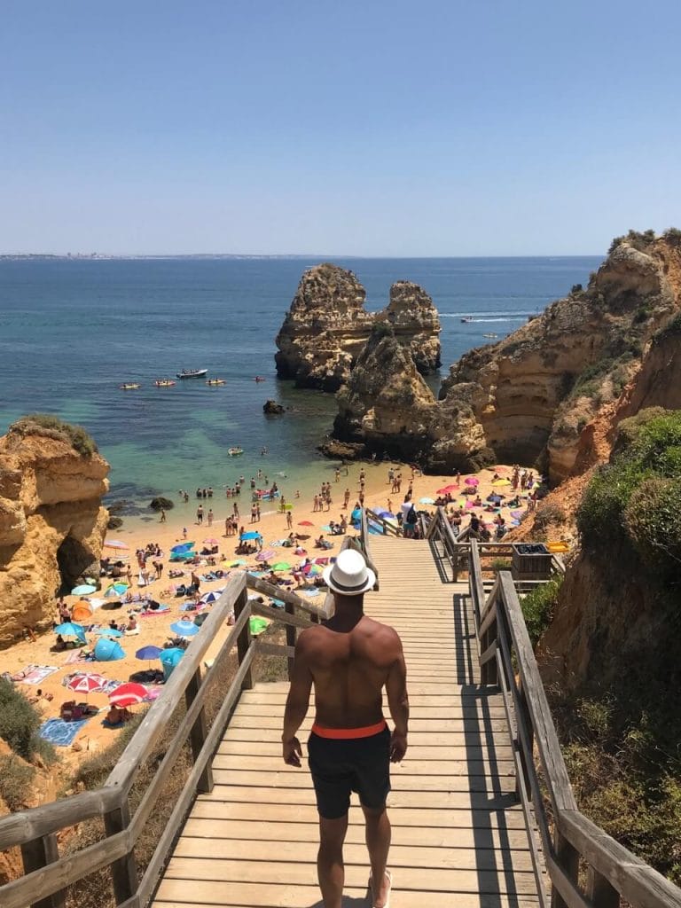 Un hombre con pantalones cortos azules y un sombrero blanco de pie en los escalones de Praia do Camilo, Lagos, con sombrillas en la arena, acantilados amarillos y kayaks en el agua