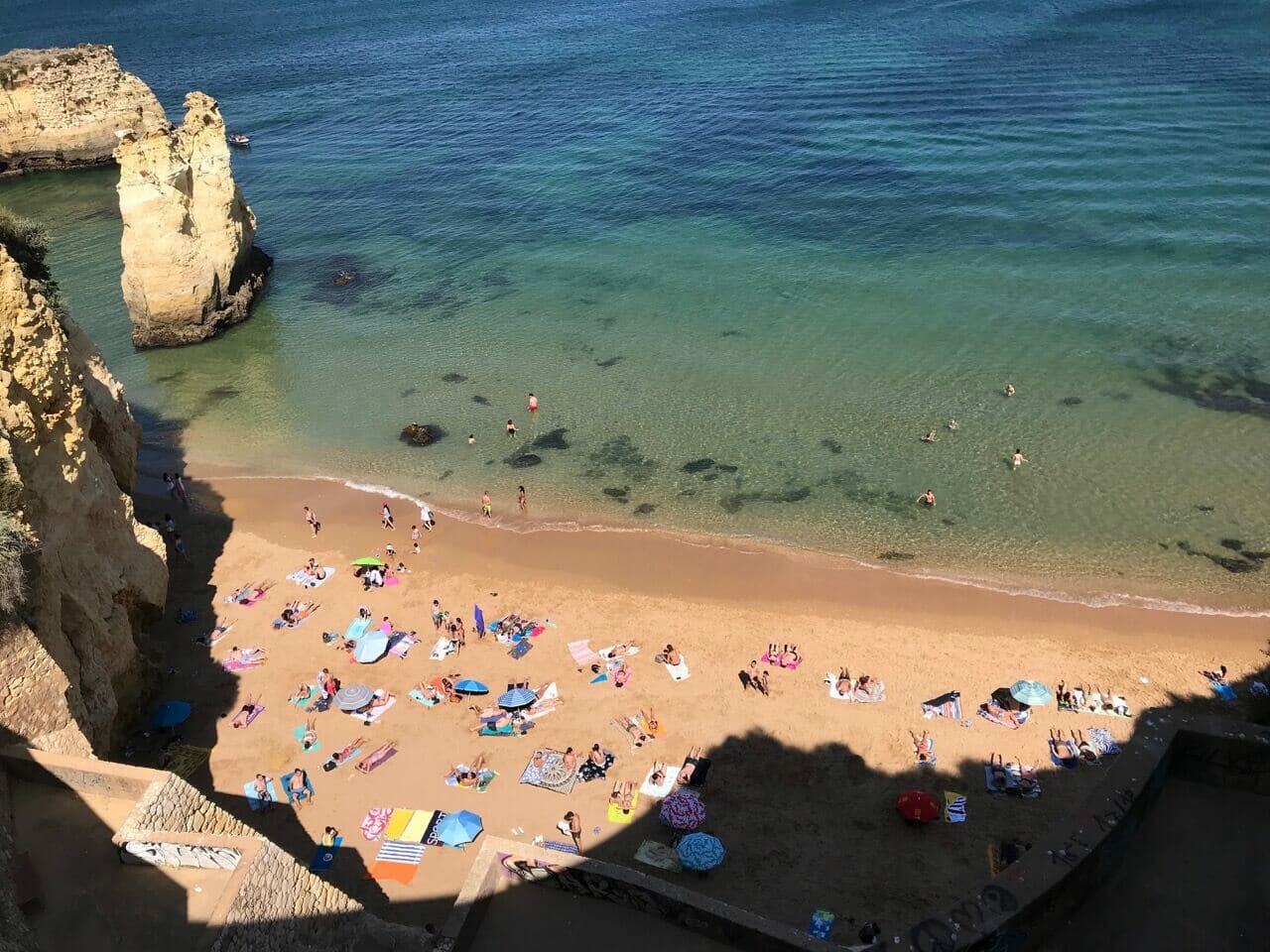 aerial view of Praia do Pinhão with many people sunbathing, others swimming in the crystalline green-blueish water some yellow cliffs